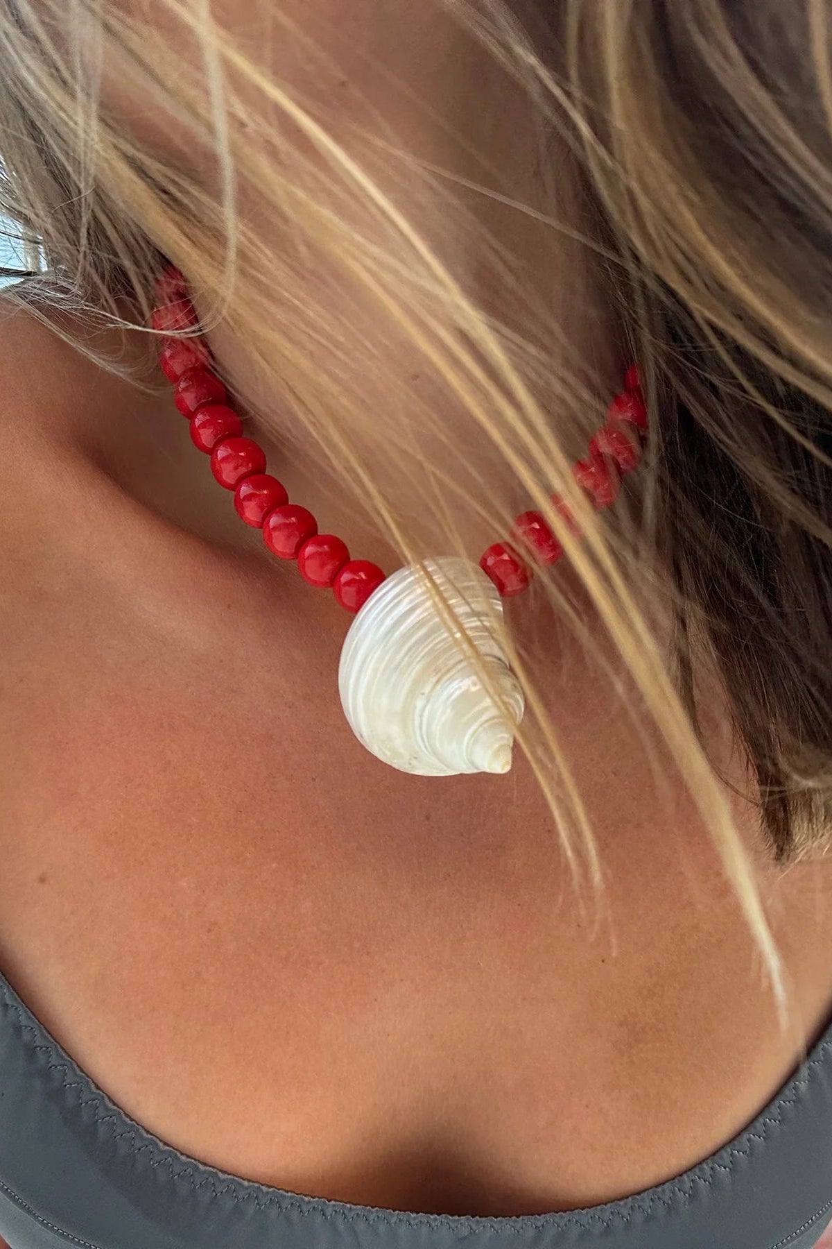 Person wearing a red beaded necklace with a shell pendant against a blurred natural background