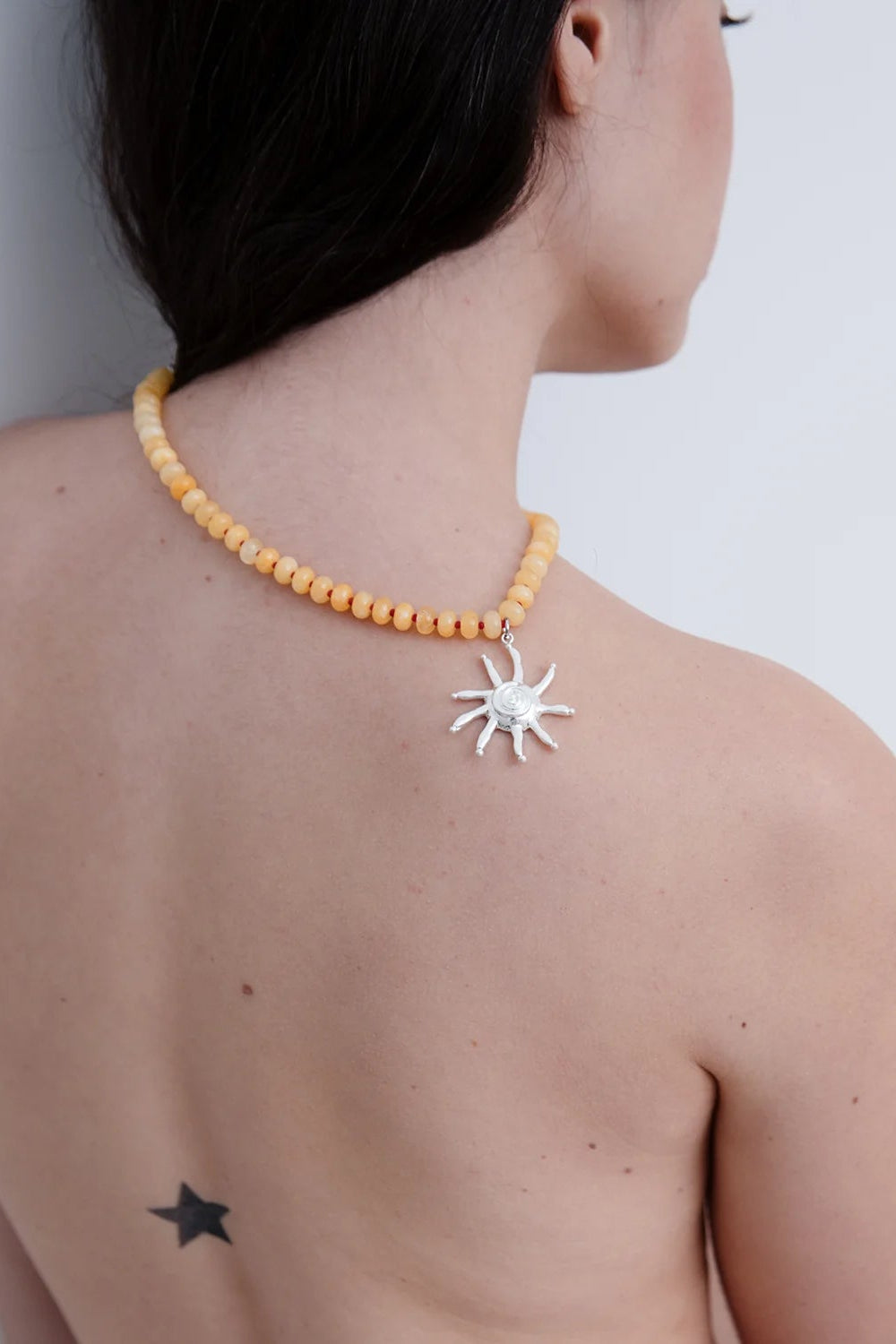 Person wearing a yellow beaded necklace with a silver sun pendant on a plain background