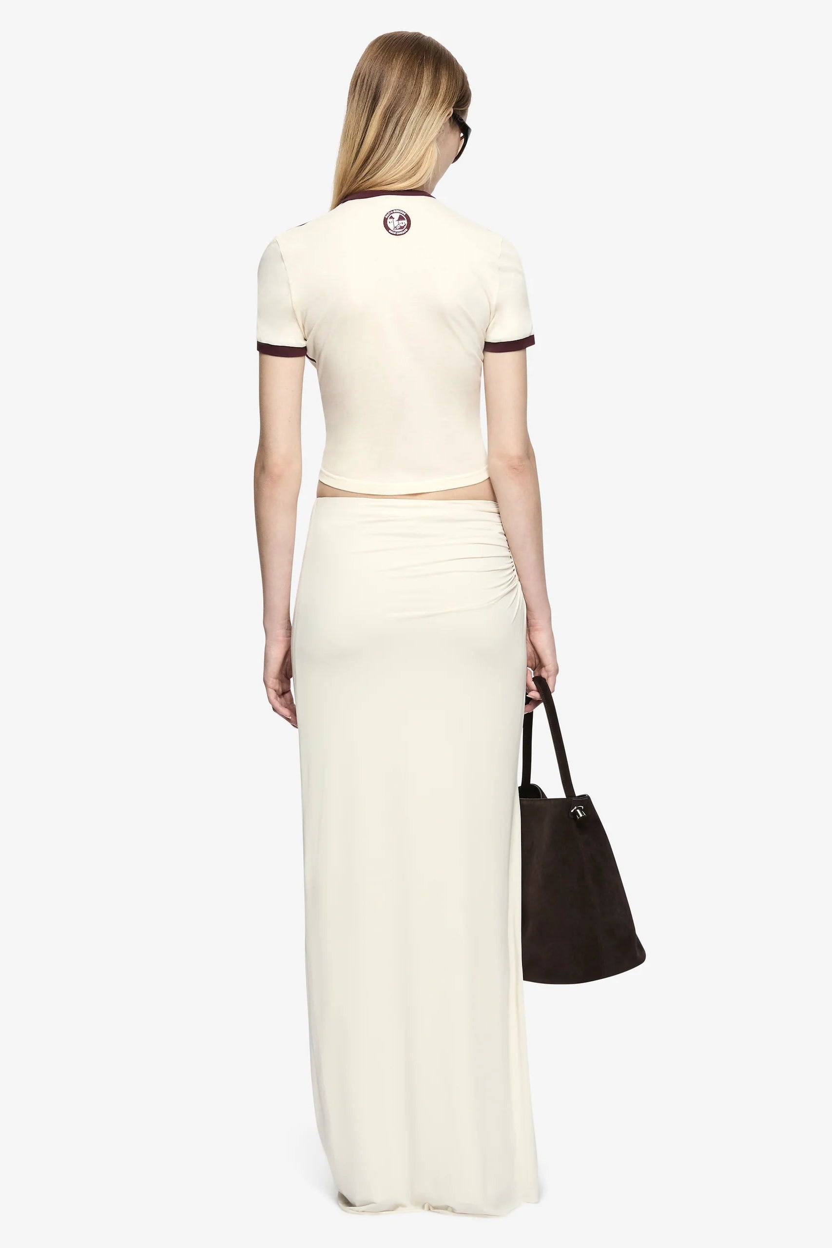 A woman shown from the back wearing a cream-colored two-piece knit set consisting of a short-sleeve crop top with dark trim and a matching long skirt. She is holding a dark brown tote bag against a solid light gray background.