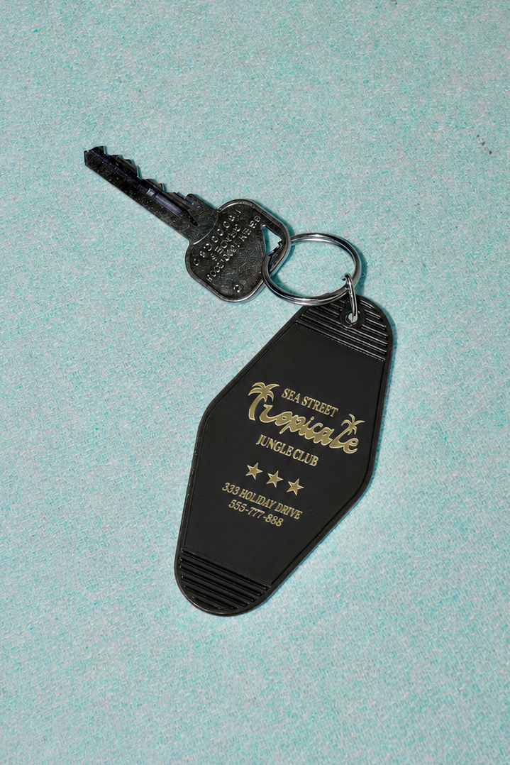 A black plastic key tag with a key attached, featuring gold and white text that reads 'The Realms of Kothath the King' and 'Royal Privilege', placed on a textured light blue surface.